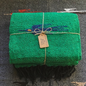 Op de afbeelding: Een gevouwen felgroene geweven deken met een donkerblauwe vogelafbeelding met een geel oog. De deken is vastgebonden met bruin touw en bevat een rechthoekig beige papieren label met de tekst SW BLANKET CO. en een afbeelding van een cactus.