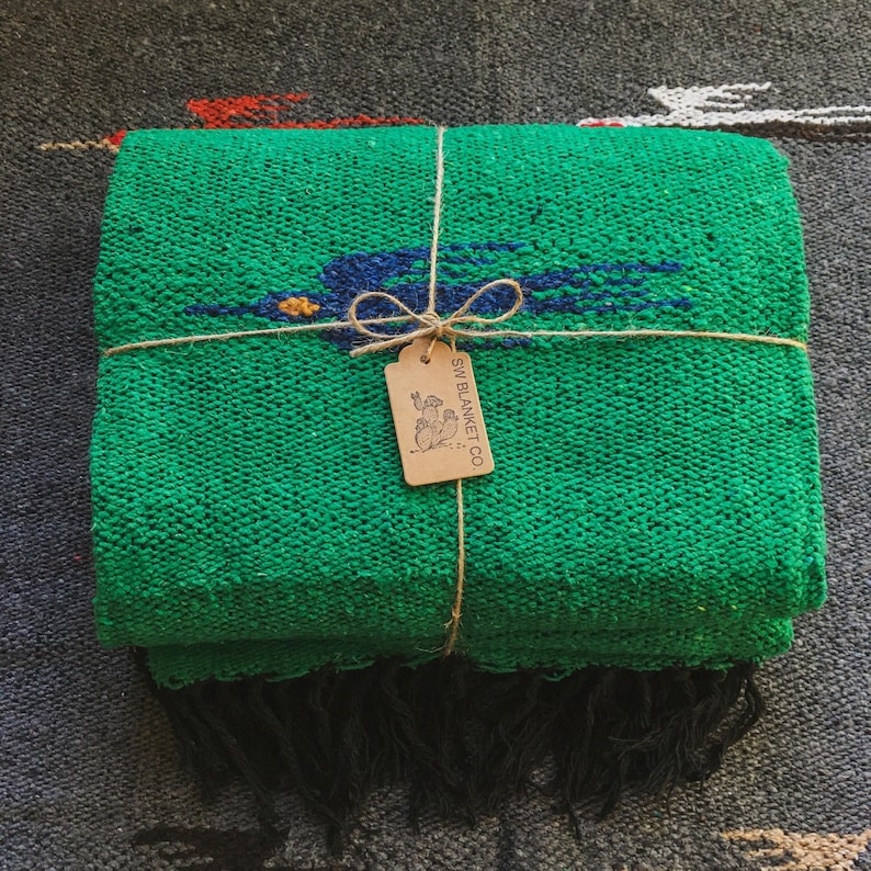 Op de afbeelding: Een gevouwen felgroene geweven deken met een donkerblauwe vogelafbeelding met een geel oog. De deken is vastgebonden met bruin touw en bevat een rechthoekig beige papieren label met de tekst SW BLANKET CO. en een afbeelding van een cactus.