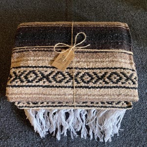 May include: A folded woven blanket featuring horizontal bands of dark brown, tan, and cream colors with a black geometric diamond pattern. The blanket is tied with twine and includes a small rectangular tag labeled SW BLANKET CO.