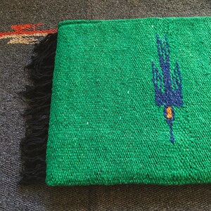 Op de afbeelding: Een levendig groen geweven textiel met een donkerblauw vogelmotief en een klein geel accent. De rechthoekige stof heeft een getextureerd, grof weefsel en is afgewerkt met een rij zwarte franjes aan &eacute;&eacute;n kant, wat een duidelijk contrast vormt.