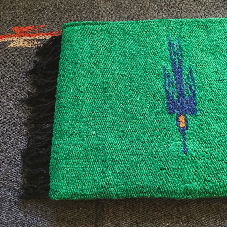 Op de afbeelding: Een levendig groen geweven textiel met een donkerblauw vogelmotief en een klein geel accent. De rechthoekige stof heeft een getextureerd, grof weefsel en is afgewerkt met een rij zwarte franjes aan &eacute;&eacute;n kant, wat een duidelijk contrast vormt.