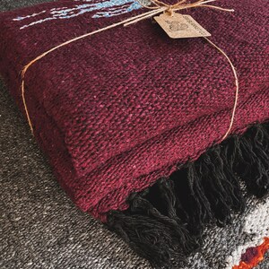 May include: A folded deep burgundy woven blanket with a thick black fringe along the edge. The blanket is tied with thin twine and features a small, rectangular tan paper tag with the text SW BLANKET CO and a graphic of a rabbit.