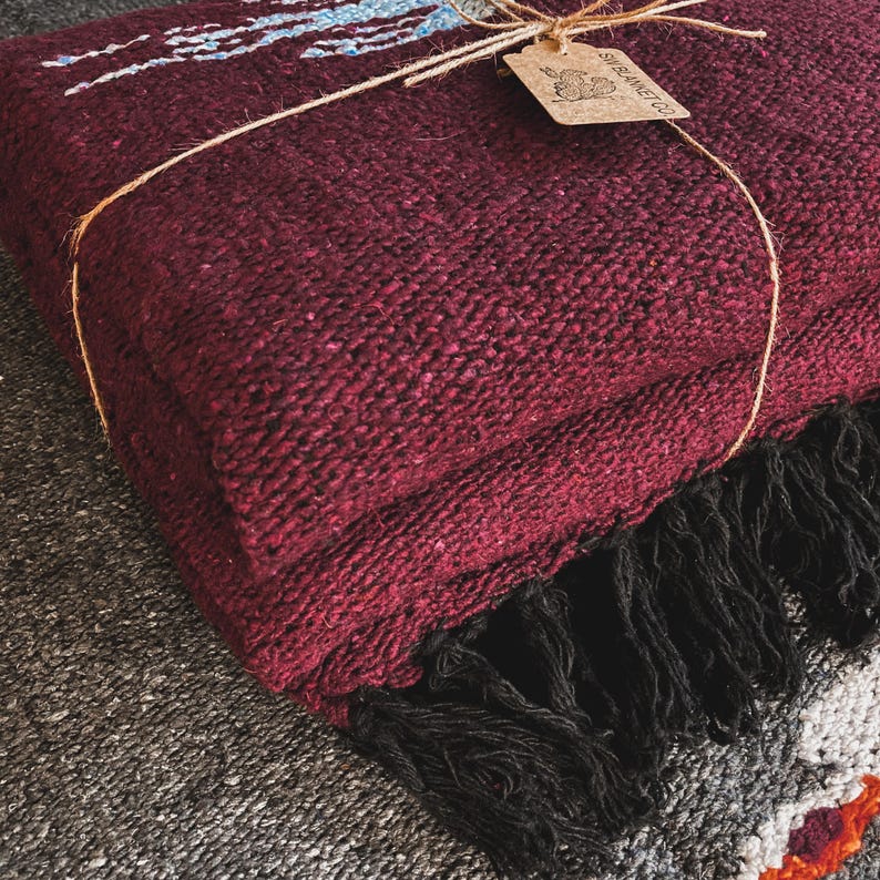 May include: A folded deep burgundy woven blanket with a thick black fringe along the edge. The blanket is tied with thin twine and features a small, rectangular tan paper tag with the text SW BLANKET CO and a graphic of a rabbit.