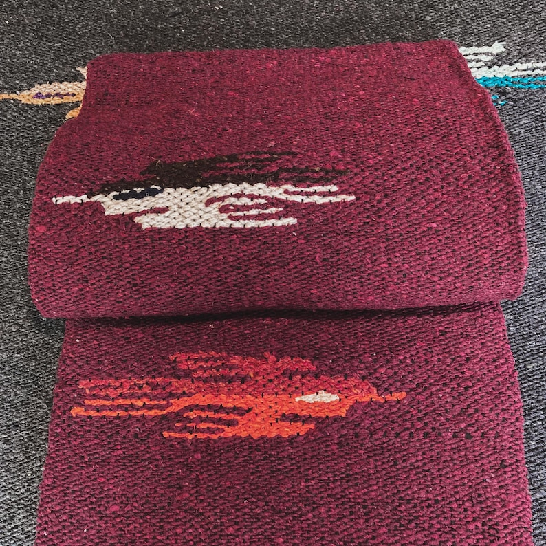 May include: A folded deep burgundy woven blanket featuring a textured surface. The top section displays a stylized bird motif in black and white, while the bottom section features a similar bird design in vibrant orange and white thread.