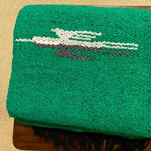Op de afbeelding: Een levendig groene geweven deken met een centrale afbeelding van een gestileerde vogel in vlucht, uitgevoerd in witte en donkergrijze draden. Het textiel heeft een getextureerd, korrelig oppervlak en is afgewerkt met donkergrijze franjes.