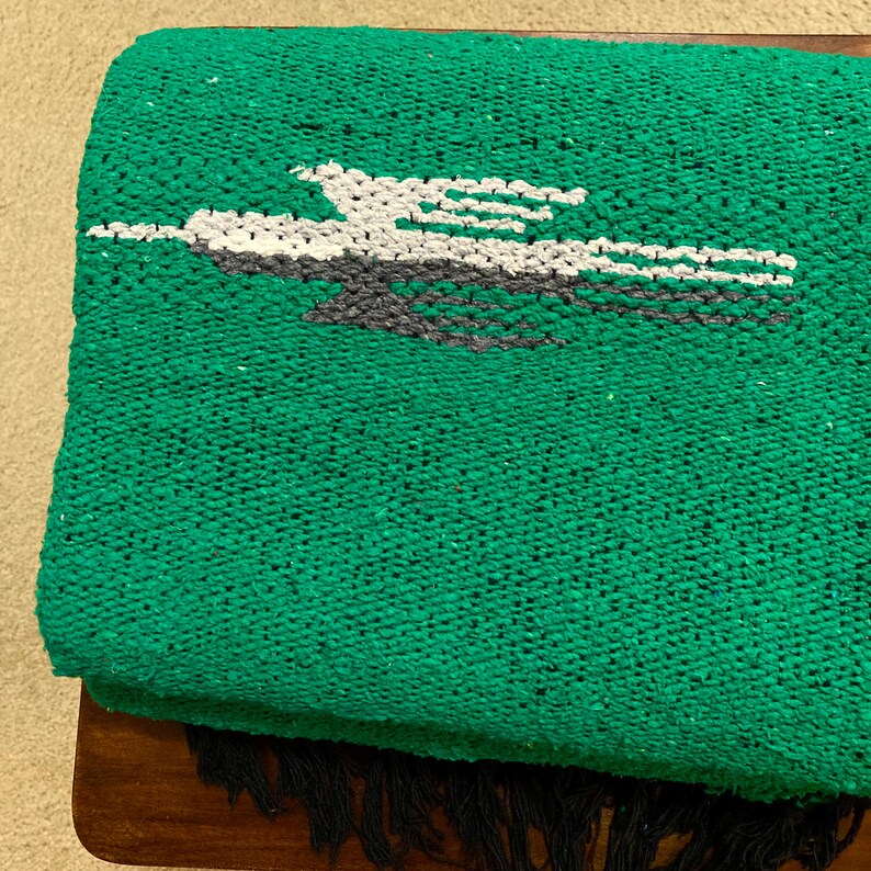 Op de afbeelding: Een levendig groene geweven deken met een centrale afbeelding van een gestileerde vogel in vlucht, uitgevoerd in witte en donkergrijze draden. Het textiel heeft een getextureerd, korrelig oppervlak en is afgewerkt met donkergrijze franjes.