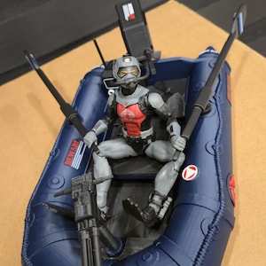 May include: An action figure in a gray and red uniform sits in a dark blue inflatable boat. The boat has black oars and a mounted weapon. The figure wears a helmet. The boat has the word 'BEWARE' printed on the side.