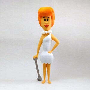 Puede incluir: Una figurita de una mujer de dibujos animados con cabello naranja, vestida con un vestido blanco y sosteniendo un palo con forma de hueso.
