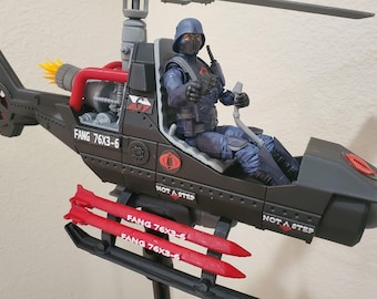 May include: A black toy helicopter with red missiles and a Cobra logo. The helicopter has the text "FANG 76X3-6" on the side and "NOT A STEP" on the bottom. A black and blue action figure is sitting in the cockpit.