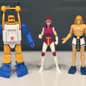 May include: Three action figures standing side by side. On the left, a yellow, white, and blue robot with a red Autobot symbol. In the center, a purple and white humanoid figure with antennae. On the right, a tan-skinned figure with blonde hair.