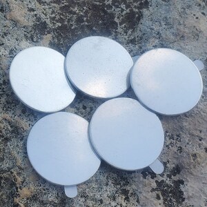 May include: Six silver-colored round metal discs with a smooth surface. The discs are arranged in a cluster on a rough, gray, textured surface.