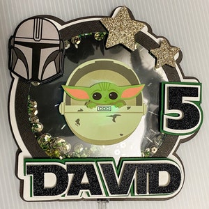 May include: A green, black, and gold cake topper featuring a character from the Star Wars franchise, a number 5, and the name "DAVID".