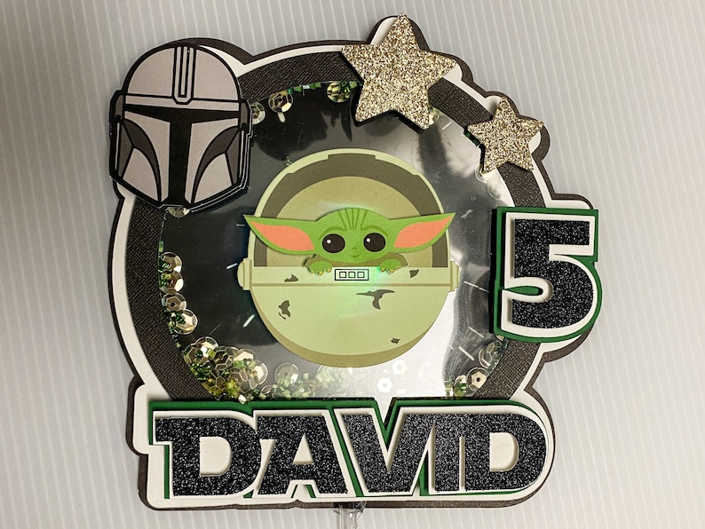 May include: A green, black, and gold cake topper featuring a character from the Star Wars franchise, a number 5, and the name "DAVID".