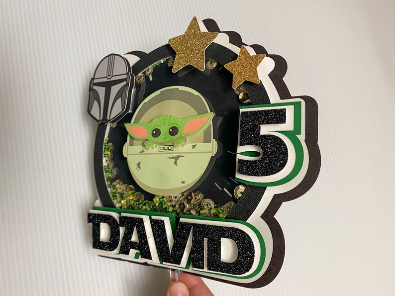 May include: A green and black cake topper featuring a character from the Star Wars franchise, a number 5, and the name "DAVID".
