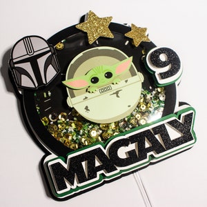 May include: A black and green cake topper with a green character from the Star Wars franchise, a black helmet, gold stars, and the name "MAGALY" in black glitter.
