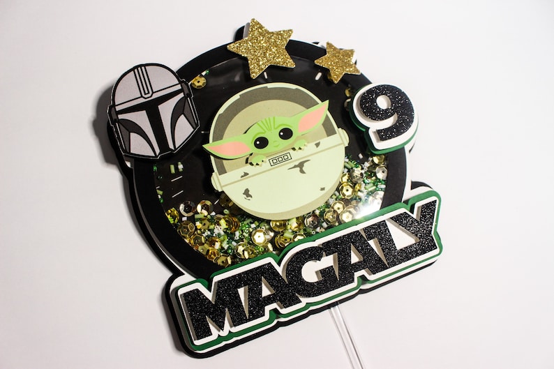 May include: A black and green cake topper with a green character from the Star Wars franchise, a black helmet, gold stars, and the name "MAGALY" in black glitter.