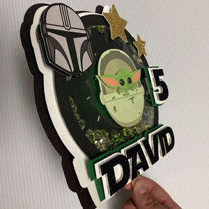 May include: A green, black, and white cake topper featuring a character from the Star Wars franchise, a number 5, and the name "DAVID".