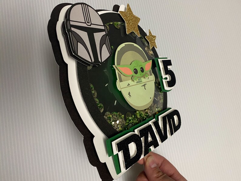 May include: A green, black, and white cake topper featuring a character from the Star Wars franchise, a number 5, and the name "DAVID".