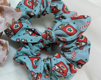 May include: A close-up of a light blue scrunchie with a repeating pattern of red coffee cups and brown dogs. The scrunchie has a bow detail and is surrounded by other scrunchies. The scrunchie is a hair accessory.