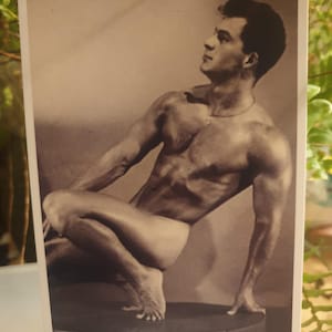 Könnte beinhalten: Sepiafarbene Vintage-Postkarte mit einer muskulösen Figur in einer sitzenden Pose, ein Bein angewinkelt und das andere ausgestreckt. Das Motiv blickt nach oben, mit einer Halskette. Das Bild ist von einem weißen Rand umgeben.