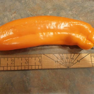 Op de afbeelding: Een feloranje paprika met een groene steel, naast een houten liniaal. De paprika is langwerpig en glad. De liniaal toont metingen in centimeters.