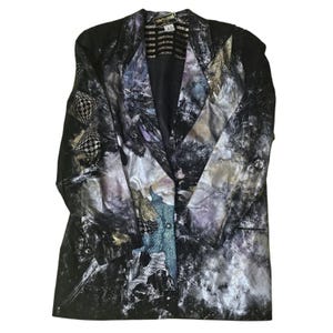 May include: A Roberto Cavalli blazer with a unique abstract design in shades of black, gray, and purple. The blazer features a notched lapel, long sleeves, and a single-button closure. The fabric has a shimmering, textured appearance.