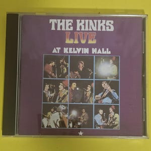 Op de afbeelding: Een paarse CD-hoes met een foto van de band The Kinks op de voorkant. De hoes heeft de titel "The Kinks Live at Kelvin Hall" erop gedrukt.