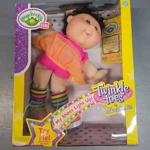 May include: Cabbage Patch Kids doll in a box. The doll wears a pink outfit with an orange overlay, striped leg warmers, and light-up shoes. The box features the text "Twinkle Toes by Skechers" and "My Shoes Light Up!"