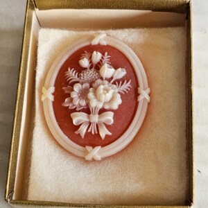 Op de afbeelding: Een witte cameo broche met een bloemmotief in een rood ovaal frame. De broche zit in een witte doos met een gouden rand.