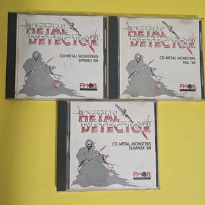 Op de afbeelding: Drie CD-hoezen met zwarte ruggen en transparante kunststof voorzijden. Elke hoes heeft een zwart-witte illustratie van een skeletfiguur die een zwaard vasthoudt. De tekst "Metal Detector" is in rood en wit op elke hoes gedrukt. De tekst "CD Metal Monsters" is onder de illustratie gedrukt, gevolgd door het seizoen van het jaar, zoals "Spring '88". De tekst "FMOB." is in de rechterbenedenhoek van elke hoes gedruckt.