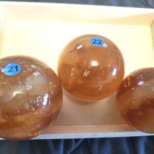 May include: Three amber-colored crystal spheres of varying sizes, labeled 21, 22, and 23, rest inside a light wooden tray. The spheres have a polished, reflective surface, and the tray has a decorative cutout design.