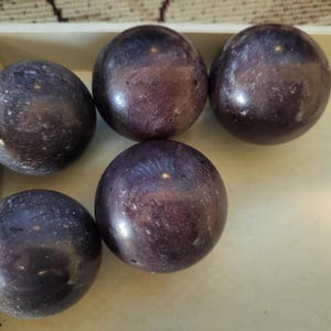 May include: Five polished, spherical, purple-colored stones with a marbled appearance are arranged in a light-colored wooden tray. The spheres have a smooth, reflective surface, and the tray sits on a patterned surface.