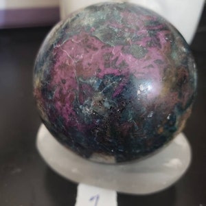 May include: A polished gemstone sphere with a mix of dark teal, purple, and gray colors. The sphere is resting on a white, oval-shaped stand. A small white tag with the number "1" is visible.