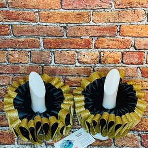 May include: Two black and gold ruffled fabric collars on white display stands. The collars are made of black satin ribbon with gold glitter trim. The collars are on a brick background. The text on the card reads "Blessing Beauties & Glitz".
