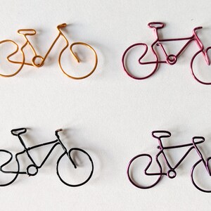 Könnte beinhalten: Vier Draht-Fahrradskulpturen in Gold, Magenta, Schwarz und Lila. Jedes Fahrrad ist aus einem einzigen Draht gefertigt und zeigt Rahmen, Räder, Sattel und Lenker. Die Fahrräder sind auf weißem Hintergrund angeordnet.