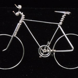 Puede incluir: Una escultura de bicicleta de alambre plateado con un marco de alambre en espiral y ruedas. La escultura est&aacute; sobre un fondo negro.