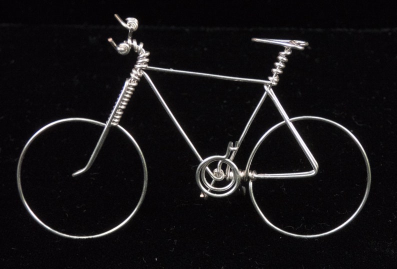 Puede incluir: Una escultura de bicicleta de alambre plateado con un marco de alambre en espiral y ruedas. La escultura est&aacute; sobre un fondo negro.