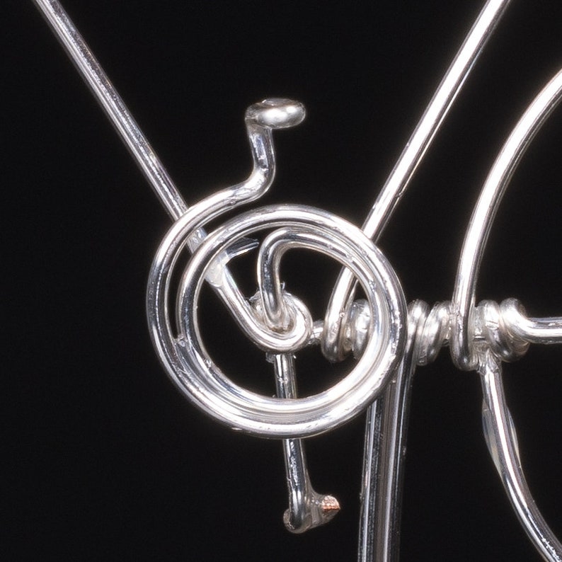 Puede incluir: Primer plano de una escultura de bicicleta de alambre plateado. La escultura est&aacute; hecha de alambre fino que se ha doblado y retorcido para crear la forma de una bicicleta. La escultura es muy detallada e intrincada.