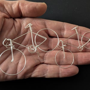 Puede incluir: Dos esculturas de bicicletas de alambre plateado, una m&aacute;s grande que la otra, se muestran en la mano de una persona. Las esculturas son detalladas y realistas, con ruedas, manillares y asientos visibles.