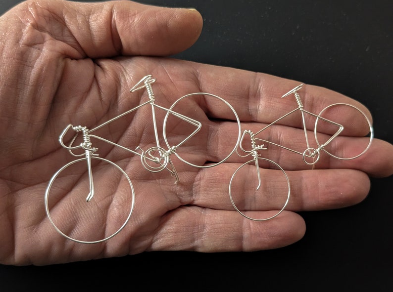 Puede incluir: Dos esculturas de bicicletas de alambre plateado, una m&aacute;s grande que la otra, se muestran en la mano de una persona. Las esculturas son detalladas y realistas, con ruedas, manillares y asientos visibles.