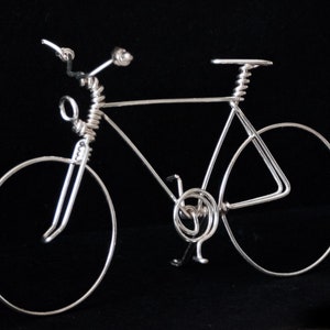 Puede incluir: Una escultura de alambre de plata de una bicicleta con un marco de alambre en espiral y ruedas. La bicicleta est&aacute; sobre un fondo negro.