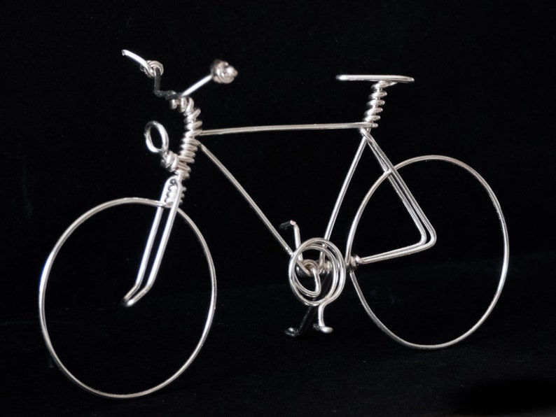 Puede incluir: Una escultura de alambre de plata de una bicicleta con un marco de alambre en espiral y ruedas. La bicicleta est&aacute; sobre un fondo negro.