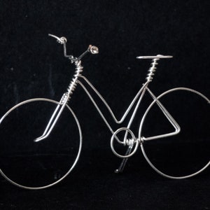 Puede incluir: Una escultura de bicicleta de alambre plateado sobre un fondo negro. La bicicleta tiene un manillar curvo, un sill&iacute;n y dos ruedas.