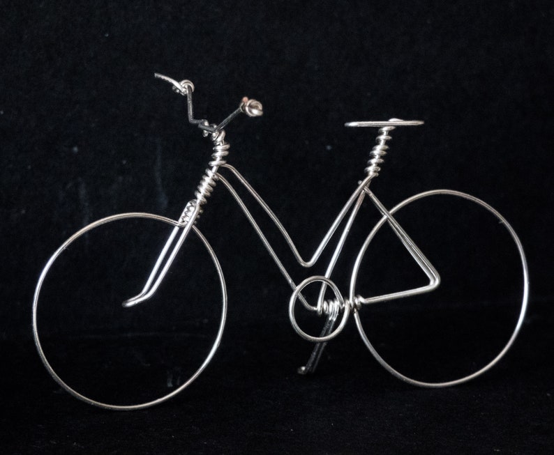 Puede incluir: Una escultura de bicicleta de alambre plateado sobre un fondo negro. La bicicleta tiene un manillar curvo, un sill&iacute;n y dos ruedas.