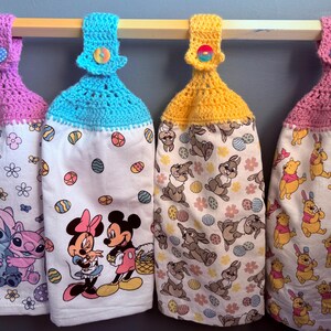 Puede incluir: Cuatro toallas de cocina de ganchillo con diferentes colores de ganchillo en la parte superior y varios dise&ntilde;os de personajes de dibujos animados. Las toallas est&aacute;n colgadas en una barra de madera con una parte superior de ganchillo rosa, azul, amarilla y rosa.