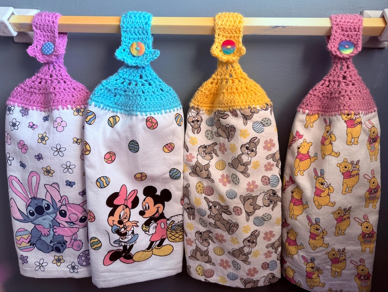 Puede incluir: Cuatro toallas de cocina de ganchillo con diferentes colores de ganchillo en la parte superior y varios dise&ntilde;os de personajes de dibujos animados. Las toallas est&aacute;n colgadas en una barra de madera con una parte superior de ganchillo rosa, azul, amarilla y rosa.