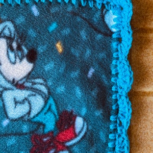 K&ouml;nnte beinhalten: Eine blaue Fleece-Decke mit einer Comicfigur, die rote Schuhe tr&auml;gt. Die Decke hat einen blauen H&auml;kelrand.