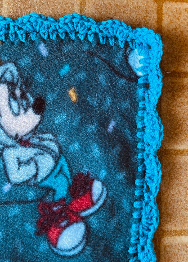 K&ouml;nnte beinhalten: Eine blaue Fleece-Decke mit einer Comicfigur, die rote Schuhe tr&auml;gt. Die Decke hat einen blauen H&auml;kelrand.