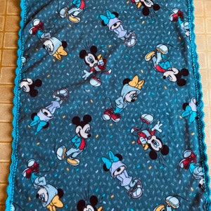 K&ouml;nnte beinhalten: Eine blaue Fleece-Decke mit einem wei&szlig;en H&auml;kelrand. Die Decke zeigt ein sich wiederholendes Muster von Mickey Mouse und Minnie Mouse Figuren in verschiedenen Posen. Der Hintergrund ist dunkelblau mit einem Konfetti-&auml;hnlichen Muster.