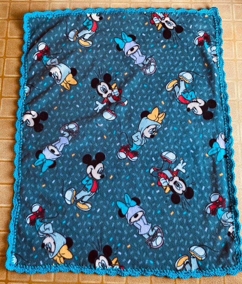 K&ouml;nnte beinhalten: Eine blaue Fleece-Decke mit einem wei&szlig;en H&auml;kelrand. Die Decke zeigt ein sich wiederholendes Muster von Mickey Mouse und Minnie Mouse Figuren in verschiedenen Posen. Der Hintergrund ist dunkelblau mit einem Konfetti-&auml;hnlichen Muster.